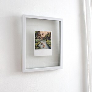 May include: A small rectangular painting in a white floating frame. The artwork depicts a cottage with a thatched roof, surrounded by greenery and flowers, with three chickens walking on a path in the foreground. The painting has a white border.