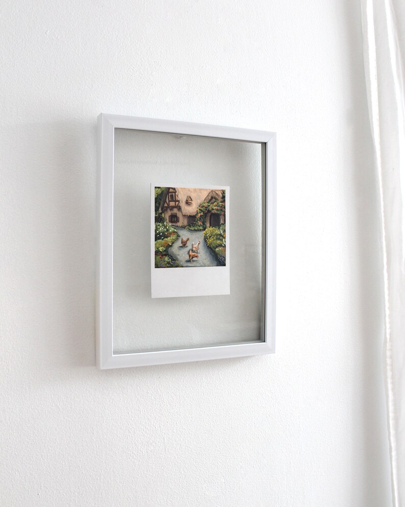 May include: A small rectangular painting in a white floating frame. The artwork depicts a cottage with a thatched roof, surrounded by greenery and flowers, with three chickens walking on a path in the foreground. The painting has a white border.
