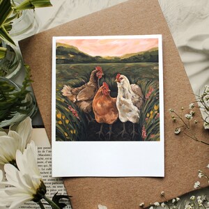 May include: A square art print featuring a painting of three chickens standing in a grassy field with yellow and pink wildflowers. The scene includes a brown chicken, a reddish-orange chicken, and a white chicken under a soft pink sunset sky.