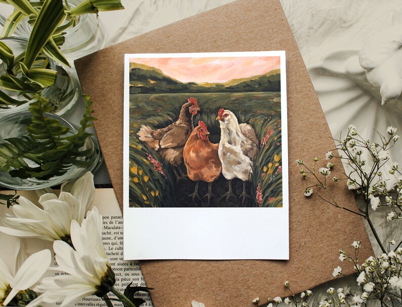 May include: A square art print featuring a painting of three chickens standing in a grassy field with yellow and pink wildflowers. The scene includes a brown chicken, a reddish-orange chicken, and a white chicken under a soft pink sunset sky.