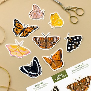 May include: A collection of die-cut butterfly paper cutouts in various colors including orange, yellow, pink, and black, alongside a pair of gold-toned metal scissors. A white paper sheet features the text "DORO MARCELLO ORIGINAL Garland version (with hole guides)" and "ORIGINALE Version guirlande (avec rep&egrave;res pour trous)".