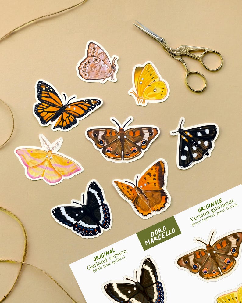 May include: A collection of die-cut butterfly paper cutouts in various colors including orange, yellow, pink, and black, alongside a pair of gold-toned metal scissors. A white paper sheet features the text "DORO MARCELLO ORIGINAL Garland version (with hole guides)" and "ORIGINALE Version guirlande (avec rep&egrave;res pour trous)".