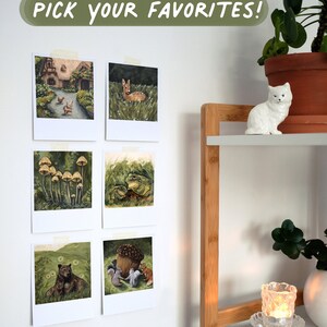 May include: Six square art prints with white borders are taped to a wall, featuring illustrations of a cottage, a deer, mushrooms, frogs, a bear, and squirrels with an acorn. The text PICK YOUR FAVORITES! is displayed in white on a green background.