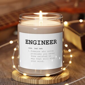 May include: A burning candle in a glass jar with a white label. The label says "ENGINEER" with a definition. The candle rests on a wooden coaster. The background includes a guitar, a book, and fairy lights.