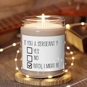 May include: A lit candle in a clear glass jar with a white label. The label reads "IS YOU A SERGEANT?" with check boxes for "YES", "NO", and "BITCH, I MIGHT BE". The candle sits on a wooden coaster, with string lights and a guitar in the background.