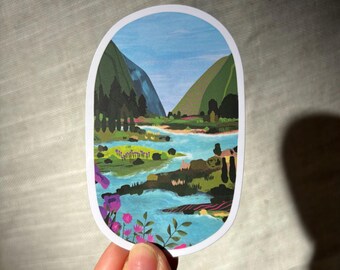 May include: Oval sticker depicting a landscape with a river winding between green hills under a blue sky. Purple flowers and foliage are at the bottom. The sticker is held by a hand.