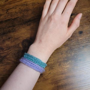 Könnte beinhalten: Zwei gehäkelte Armbänder am Handgelenk einer Person. Das obere Armband ist türkis und das untere Armband ist lavendelfarben.