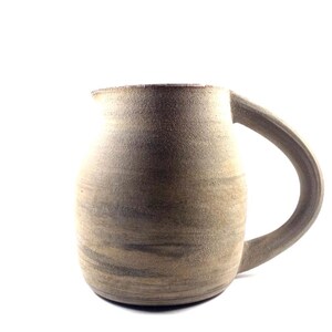 Peut inclure: Une tasse en grès faite à la main avec une grande anse incurvée. La tasse a un corps arrondi et un aspect texturé et terreux avec des nuances de gris et de brun. La tasse est conçue pour contenir des boissons.