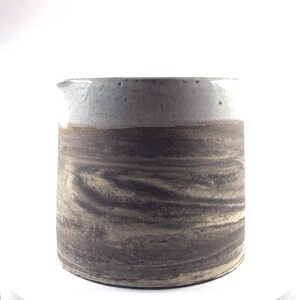 Peut inclure: Une cruche en céramique faite à la main avec un design marbré unique. Le bord supérieur est de couleur gris clair, avec une bande marron le séparant du corps plus foncé, tourbillonnant gris et beige. La cruche a un petit bec verseur.