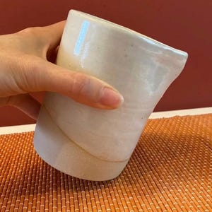 Op de afbeelding: Handgemaakte, gebroken witte keramische kan met een uniek, asymmetrisch ontwerp. De kan heeft een licht gebogen lichaam en een tuit om gemakkelijk te schenken. Het oppervlak heeft een subtiele, getextureerde uitstraling. De kan wordt boven een oranje bamboemat gehouden.