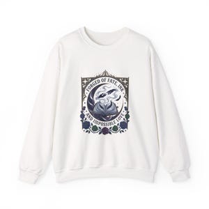 Peut inclure: Sweat-shirt blanc avec un motif graphique représentant un livre ouvert, de la fumée et le texte "FORGED OF FATE, INK, AND IMPOSSIBLE LOVE". Le motif comprend un croissant de lune et des roses bleues, violettes et vertes.