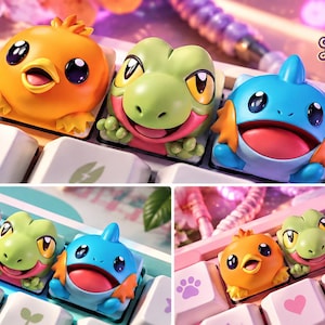 May include: Three custom artisan mechanical keyboard keycaps featuring sculpted, colourful characters: an orange chick, a green reptile, and a blue creature with orange fins. The keycaps are set on a white keyboard with pastel icons.