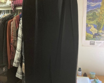 Pode incluir: Uma saia midi preta lisa de cintura subida com silhueta reta, fecho de correr centrado nas costas e uma racha vertical na bainha inferior. A saia é construída com costuras verticais subtis que vão desde a cintura até à bainha da saia.