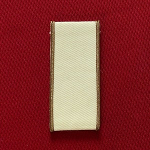 Könnte beinhalten: Ein weißes Stofflabel mit goldener Umrandung. Das Label ist rechteckig und hat eine glatte Oberfläche.