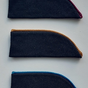 May include: Three navy blue fabric patches with a curved edge, each trimmed with a different color of rope: burgundy, gold, and blue. The patches are rectangular with a curved edge.