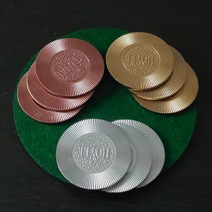 Puede incluir: Fichas de casino en cobre, oro y plata, cada una con un diseño radial y la palabra "HOTEL". Las fichas están dispuestas sobre un círculo de fieltro verde. Para juegos de azar.