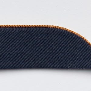 May include: A navy blue fabric patch with a gold-colored, twisted rope trim along the curved edge. The patch has a rectangular shape with one rounded corner. The fabric has a subtle diagonal weave.