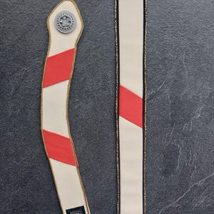 Puede incluir: Dos correas de tela color blanco roto con rayas rojas diagonales y ribete metálico dorado. La correa izquierda es curva con un emblema gris en forma de engranaje en la parte superior y un pequeño componente rectangular negro en la base.