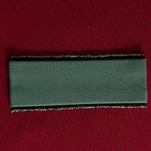 May include: A gray fabric patch with a black border and gold trim. The patch is rectangular and has a smooth surface.
