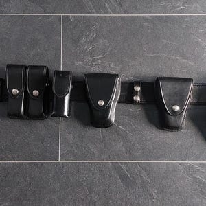 Può includere: Una cintura di servizio in pelle nera con fibbia in metallo argentato, due tasche rettangolari, una custodia verticale sottile, due supporti per attrezzatura sagomati e una fondina, il tutto fissato con bottoni a pressione in metallo.