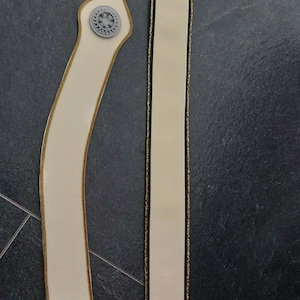 May include: Two off-white fabric strips with gold trim. The left strip is curved, features a circular grey emblem at the top, a rectangular silver buckle at the bottom, and a separate small silver metal clasp. The right strip is straight.