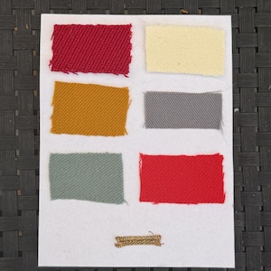 May include: A white rectangular card featuring six rectangular fabric swatches arranged in two columns. The colors include red, cream, mustard yellow, gray, sage green, and a second red swatch, with a small gold metallic thread piece at the bottom.