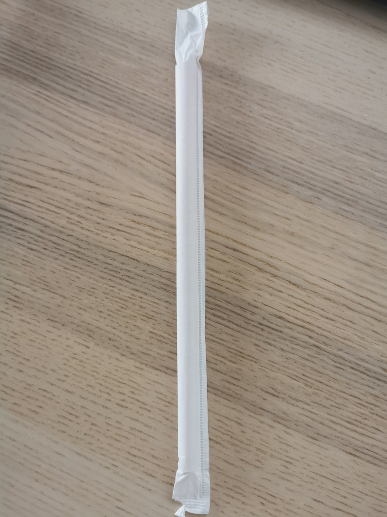 May include: A white paper drinking straw, wrapped in a white paper wrapper, is lying flat on a wooden surface.