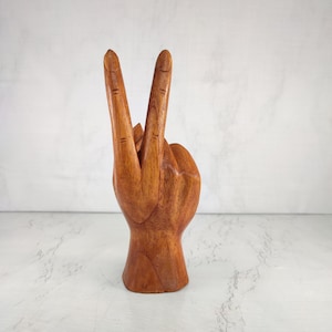 May include: A wooden hand sculpture with two fingers raised in a peace sign. The hand is carved from a light brown wood and has a smooth finish.