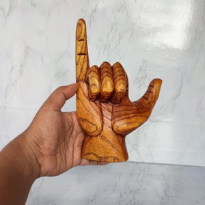 May include: A wooden hand sculpture in the shape of the "hang loose" hand gesture. The hand is carved from a light brown wood with a reddish-brown grain.