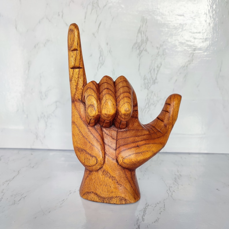 May include: A wooden hand sculpture in the shape of the "hang loose" gesture, with the index finger and pinky extended and the other fingers curled. The sculpture is made of light brown wood with a natural grain pattern.