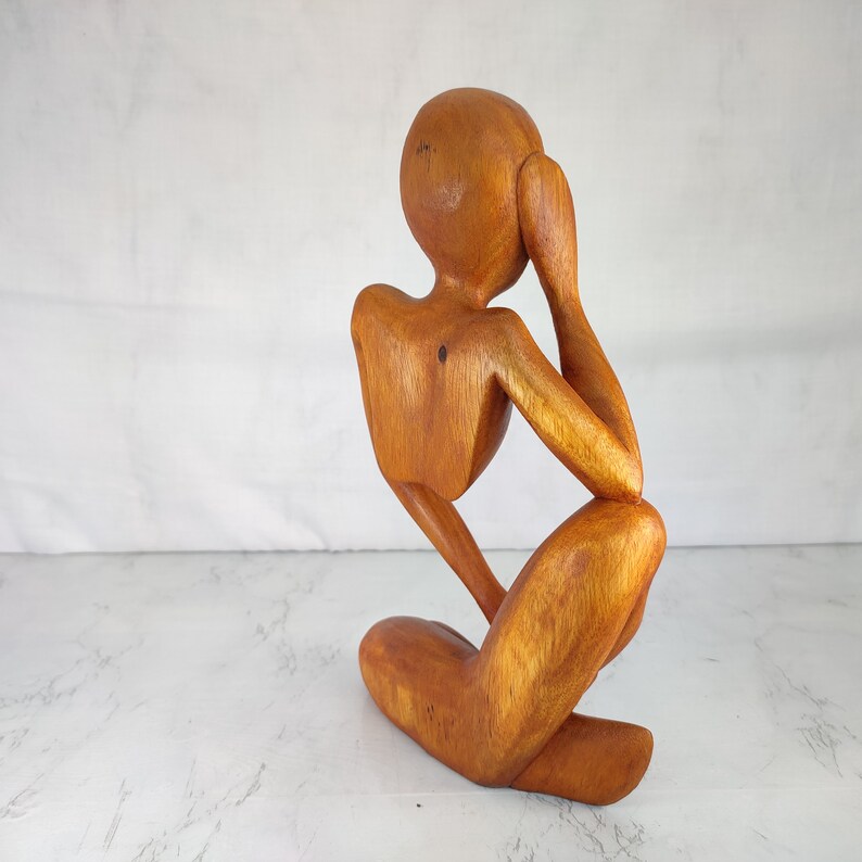 May include: A wooden sculpture of a person sitting with their head in their hand, carved in a minimalist style. The sculpture is made of light brown wood and has a smooth finish.