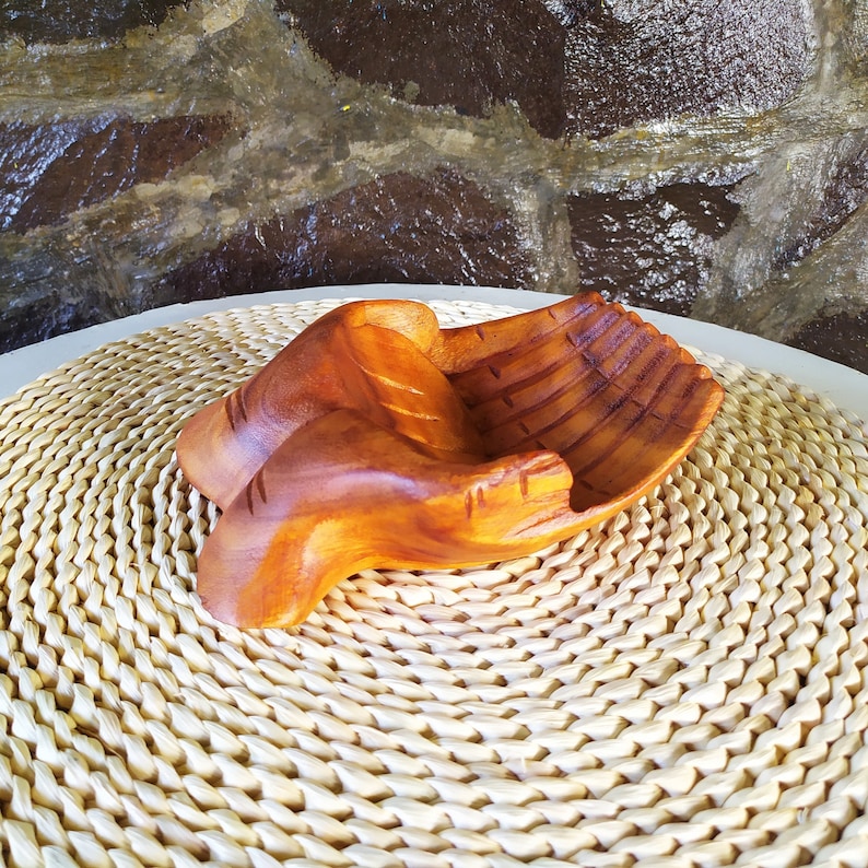 Pu&ograve; includere: Una ciotola di legno scolpita a forma di due mani giunte. La ciotola &egrave; di colore marrone chiaro e ha una finitura liscia e lucida.