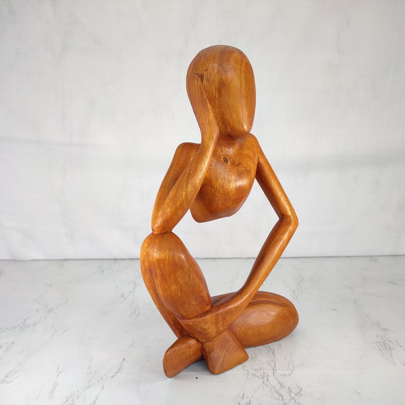 May include: A wooden sculpture of a person sitting with their chin resting on their hand, in a contemplative pose. The sculpture is made of a light brown wood and has a smooth, polished finish.
