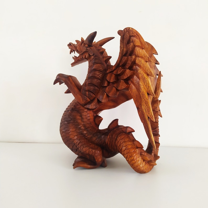 May include: A wooden dragon sculpture with detailed wings and scales. The dragon is carved from a single piece of wood and has a natural brown colour.