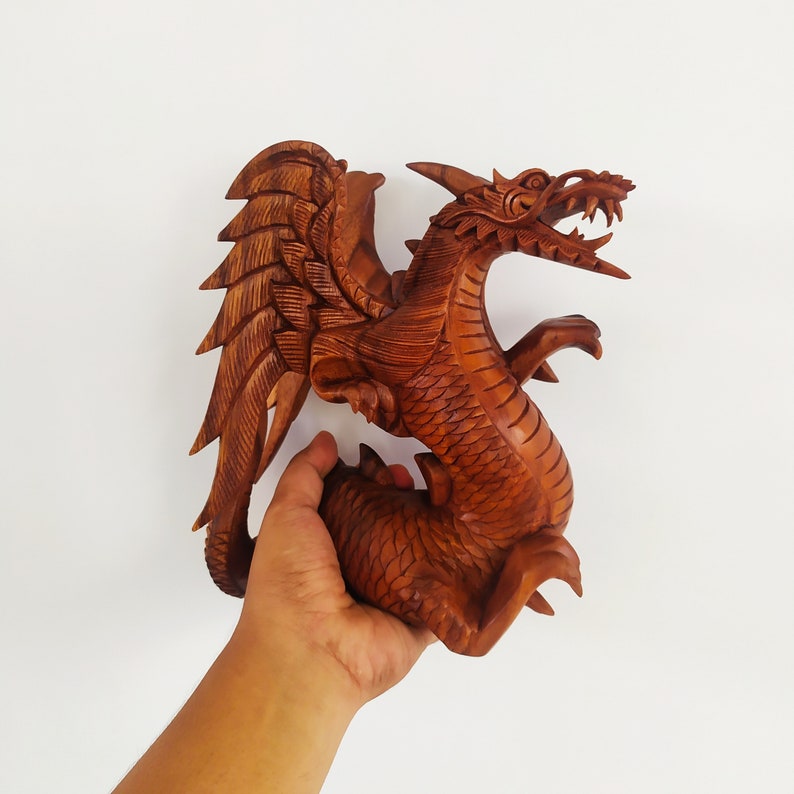 May include: A hand holds a wooden carving of a dragon with outstretched wings. The dragon is brown and has detailed scales and claws.