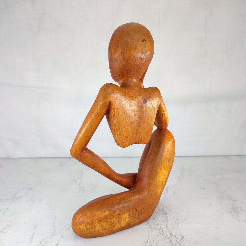 May include: A wooden sculpture of a person sitting with their head tilted down. The sculpture is made of light brown wood and has a smooth, polished finish.