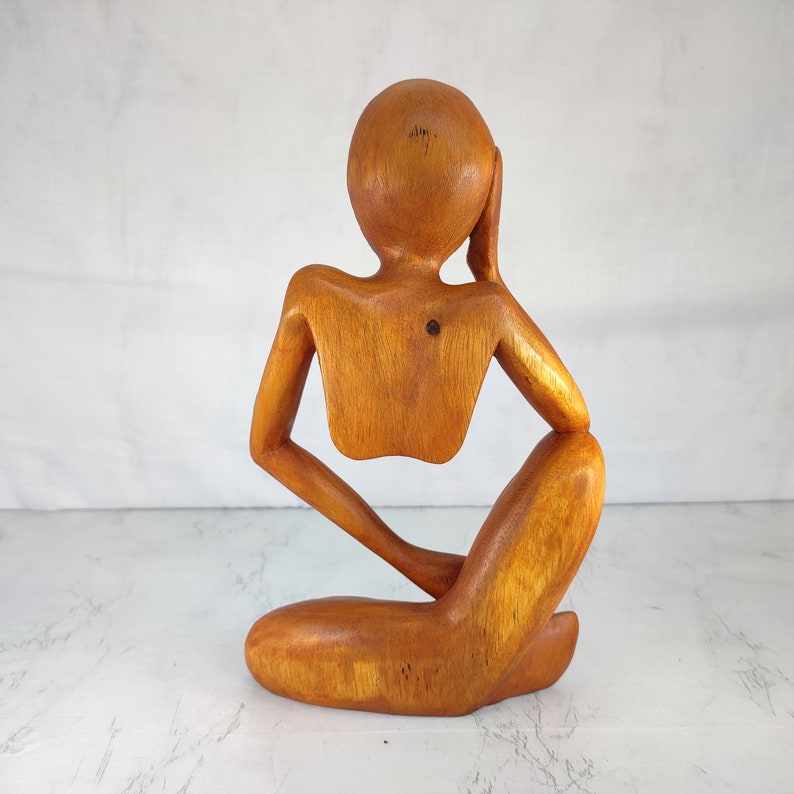 May include: Wooden sculpture of a person sitting with their head resting on their hand. The sculpture is made of light brown wood and has a smooth finish.