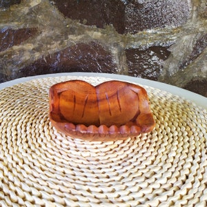 Pu&ograve; includere: Una ciotola di legno con un design unico intagliato a mano. La ciotola &egrave; marrone e ha una finitura in legno naturale. &Egrave; appoggiata su un tappetino bianco intrecciato.