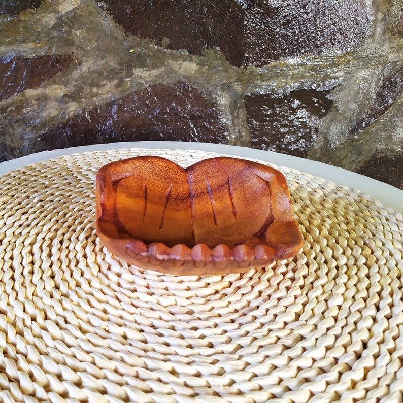 Pu&ograve; includere: Una ciotola di legno con un design unico intagliato a mano. La ciotola &egrave; marrone e ha una finitura in legno naturale. &Egrave; appoggiata su un tappetino bianco intrecciato.