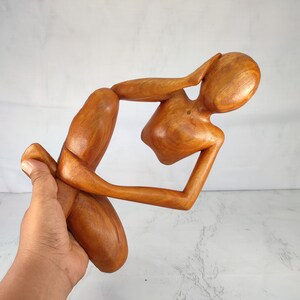 May include: A wooden sculpture of a person in a seated position with their head resting on their hand. The sculpture is made of a light brown wood and has a smooth finish.