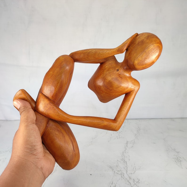 May include: A wooden sculpture of a person in a seated position with their head resting on their hand. The sculpture is made of a light brown wood and has a smooth finish.