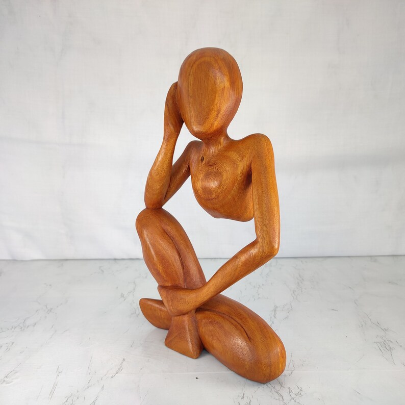 May include: A wooden sculpture of a person sitting with their head resting on their hand. The sculpture is made of a light brown wood and has a smooth finish.
