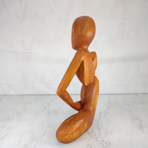 May include: A wooden sculpture of a person in a seated pose. The sculpture is made of light brown wood and has a smooth, polished finish.