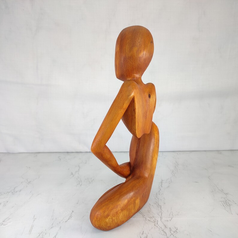 May include: A wooden sculpture of a person in a seated pose. The sculpture is made of light brown wood and has a smooth, polished finish.