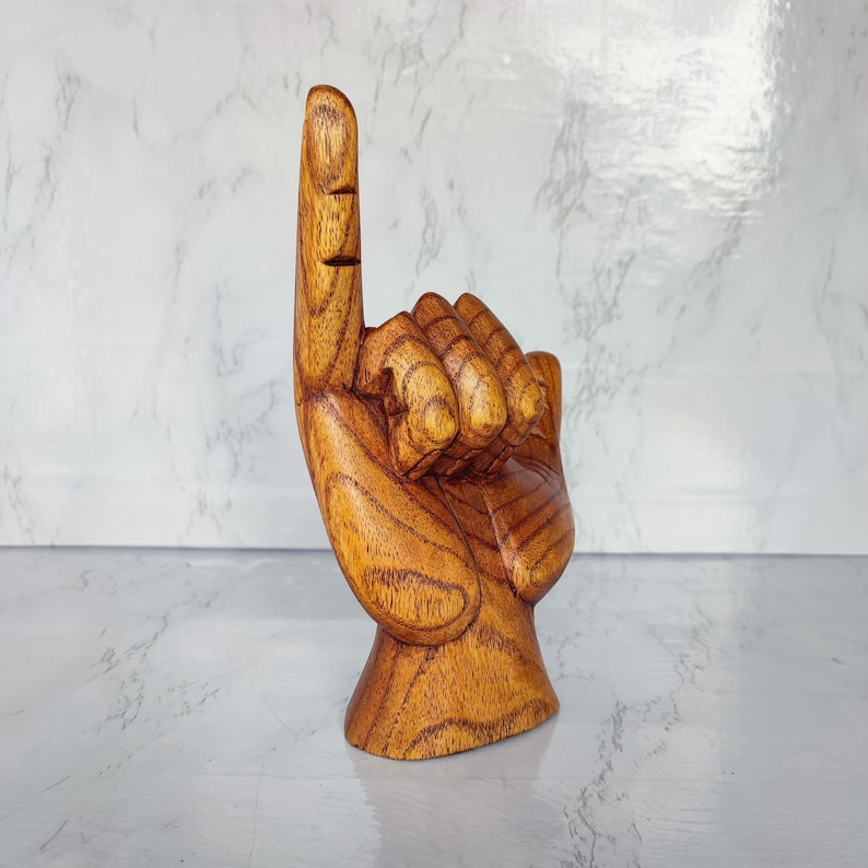 May include: A wooden hand sculpture with one finger pointing upwards. The hand is carved in a realistic style and has a warm brown color.