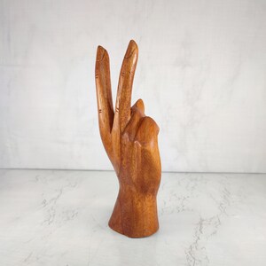 May include: A wooden hand sculpture with two fingers raised in a peace sign. The hand is carved from a light brown wood and has a smooth finish.