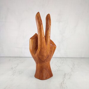 May include: A wooden hand sculpture with two fingers raised in a peace sign gesture. The hand is carved from a light brown wood and has a smooth, polished finish.
