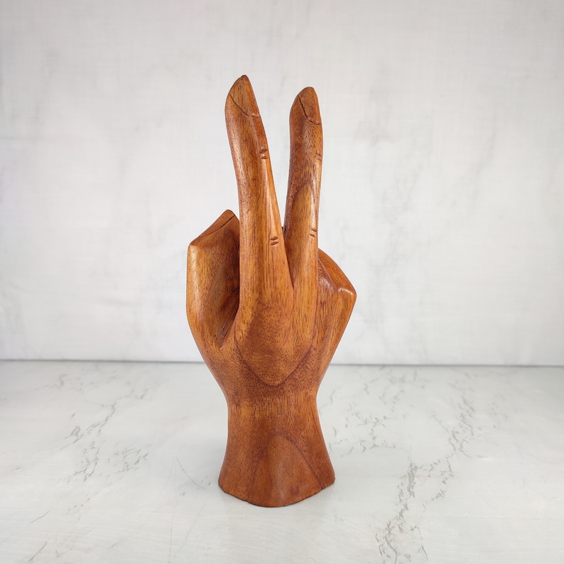 May include: A wooden hand sculpture with two fingers raised in a peace sign gesture. The hand is carved from a light brown wood and has a smooth, polished finish.
