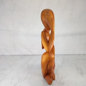 May include: A wooden sculpture of a person sitting with their head in their hands. The sculpture is made of a light brown wood and has a smooth finish.