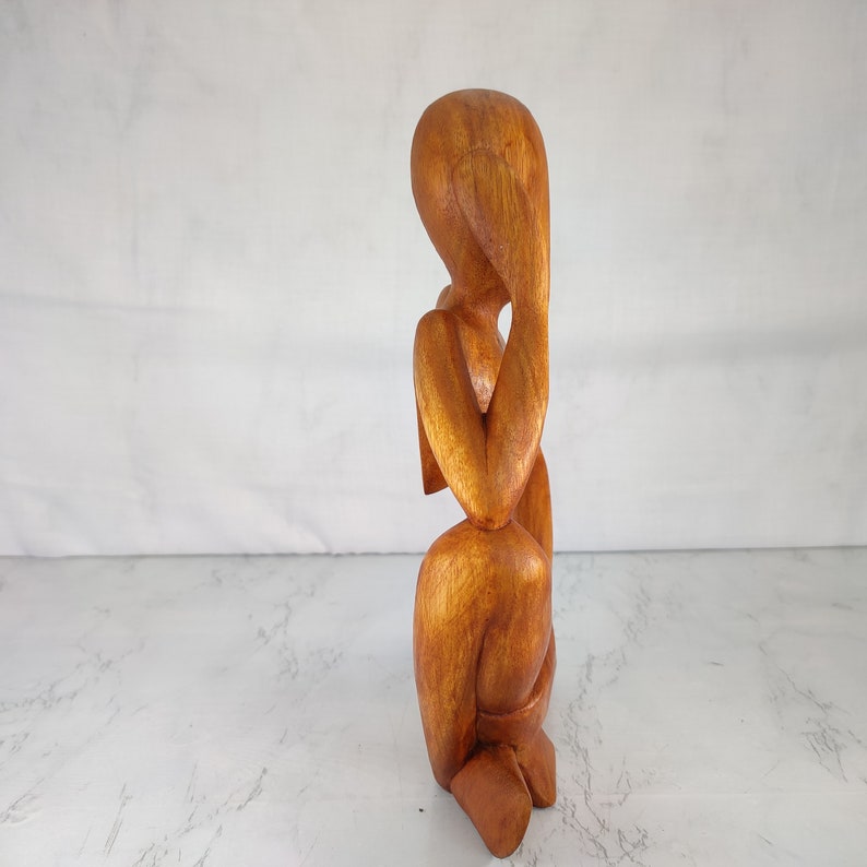May include: A wooden sculpture of a person sitting with their head in their hands. The sculpture is made of a light brown wood and has a smooth finish.